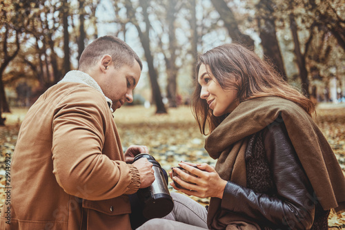 Smiling happy couple is drinking hot drink from thermos while sitting at autumn park. © Fxquadro