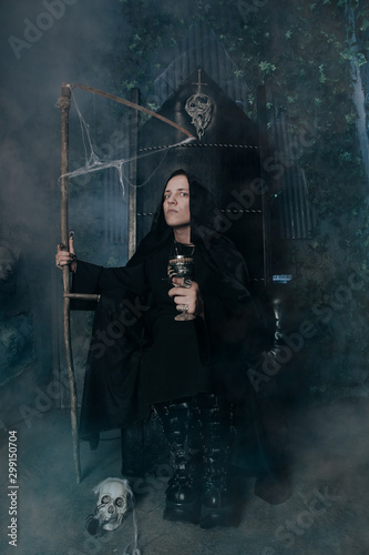 young very angry man with a caucasian face is dressed in a black robe and leather big boots and sits with scythe on scary vampire throne in the dark at Halloween night.