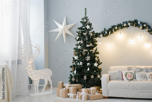 A beautiful Christmas tree stands in the interior of the living room with a sofa and a deer with lights