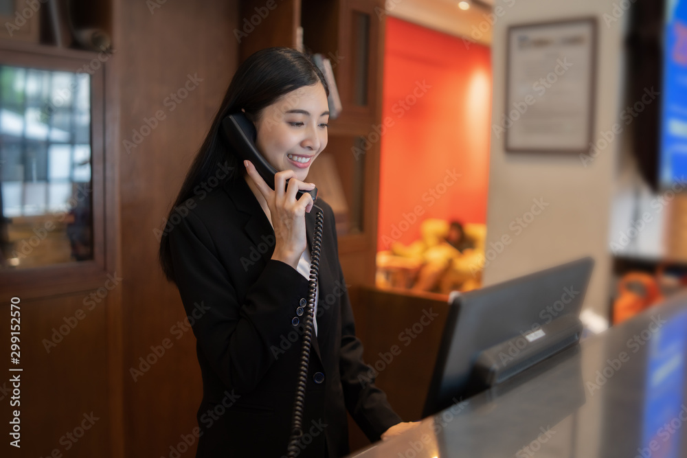 Welcome to the hotel,Happy young Asian woman hotel receptionist worker smiling standing,she taking  telephone call at a Modern luxury reception counter waiting for guests getting key card in hotel - obrazy, fototapety, plakaty 