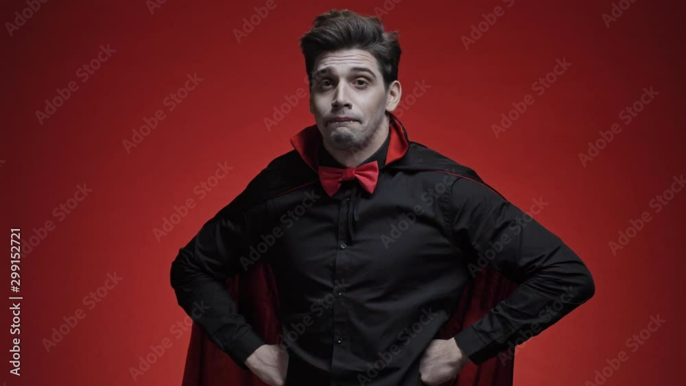 Vampire man with fangs in black halloween costume becoming confused and shrugging his shoulders isolated over red wall Vampire man with fangs in black halloween costume becoming confused and shrugging his shoulders isolated over red wall