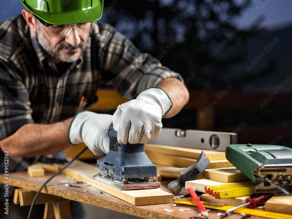 Adult carpenter craftsman wearing helmet and leather protective gloves, with the electric sander smoothes a wooden table. Construction industry, housework do it yourself. Safety at work. Adult carpenter craftsman wearing helmet and leather protective gloves, with the electric sander smoothes a wooden table. Construction industry, housework do it yourself. Safety at work.