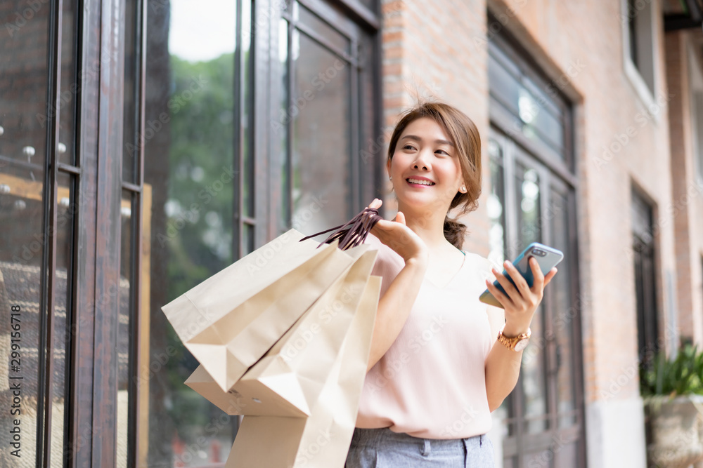 Fototapeta premium Smiling Asian Thai girl holding gift in shopping bags She Compares prices on mobile smartphone looking for clothes in a dress shop - Concepts of relaxing lifestyle,shopping online technology in city