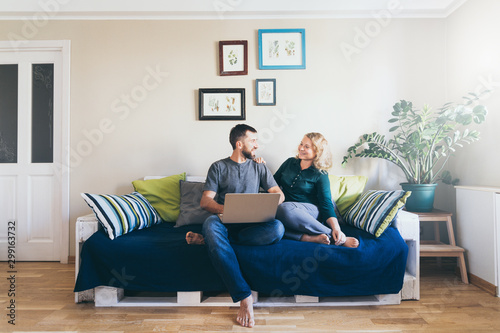 Young couple doing shoppings online on the sofa at home with laptop, looking at each other and smiling