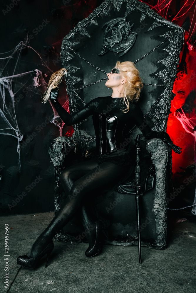 Naklejka premium Gothic girl with scary makeup on a huge throne ready for Halloween party. Young blonde woman with little bird on a black chair.
