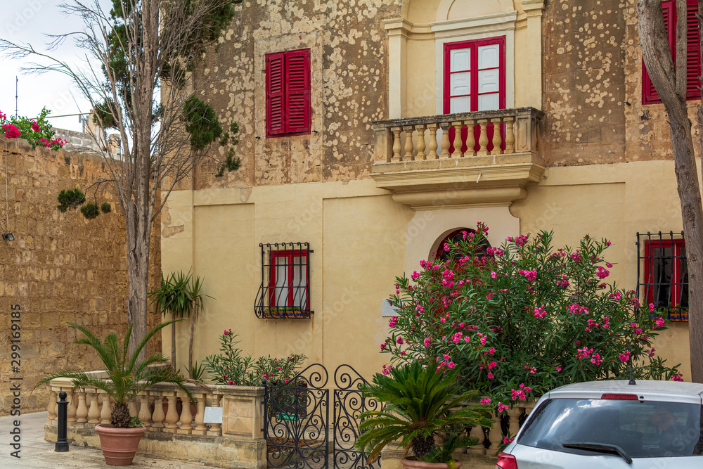Naklejka premium Picturesque house in Mdina, Malta, with brightly red door and windows' frames and shutters, and with lush Nerium oleander bush growing near the fence door gate.