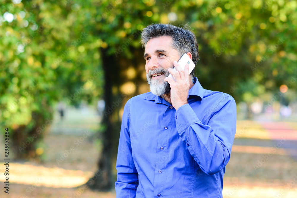 Portrait of modern senior man speaking by phone outdoors. Happy bearded senior man talking by phone. Cheerful senior businessman talking by smartphone