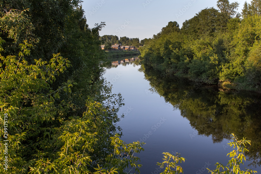 Calm river with overgrown banks, in the distance on the shore are visible wooden houses
