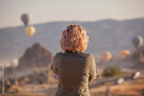 woman is watching on scenery view with rising balloons on sunrise. Girl in gorgeous pink long dress stay on hill looking at large number of air balls. Fabulous Cappadocia mountains landscapes Turkey