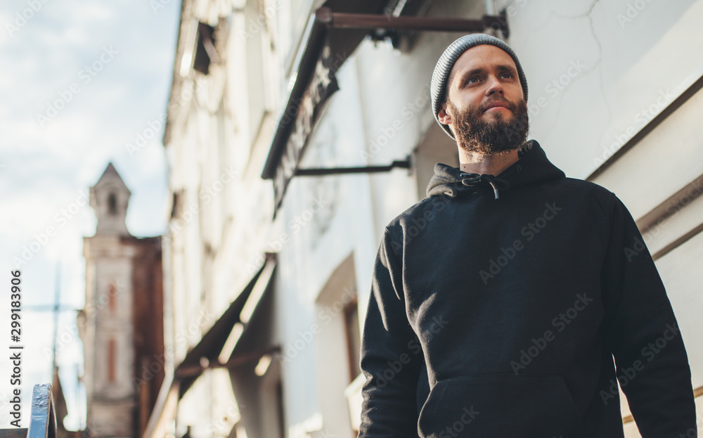 City portrait of handsome hipster man with beard wearing black blank hoodie or hoody and hat with space for your logo or design. Mockup for print - obrazy, fototapety, plakaty 
