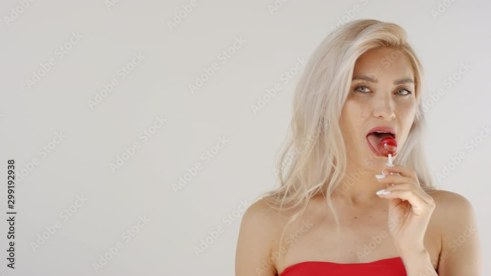 Chest-up shot of blonde Caucasian girl with long hair and bare shoulders posing in studio, sucking and licking lollipop while staring provocatively at camera Chest-up shot of blonde Caucasian girl with long hair and bare shoulders posing in studio, sucking and licking lollipop while staring provocatively at camera