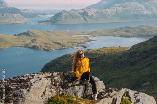 Girl in yellow jacket stitting on stone in mountains and looking to camera