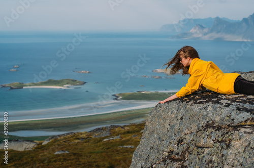 Girl in yellow jacket lies on stone in on the edge of rocks in mountains and looking to fjord