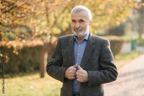 Handsome elderly man putting on a gray jacket. Old gray-haired bearded man walk in the autumn park. Yellow background