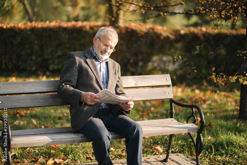 Handsome grandfather with a beautiful beard in a gray jacket sits on a bench in the park and reads a newspaper. Senior gray-haired man in glasses Handsome grandfather with a beautiful beard in a gray jacket sits on a bench in the park and reads a newspaper. Senior gray-haired man in glasses