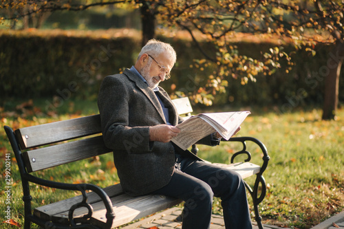 Handsome grandfather with a beautiful beard in a gray jacket sits on a bench in the park and reads a newspaper. Senior gray-haired man in glasses