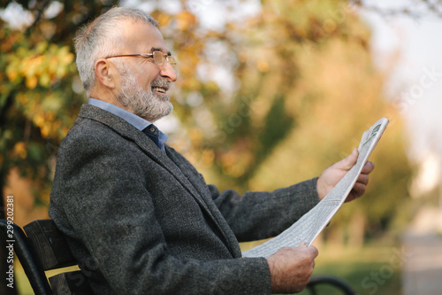 Bearded elderly man in glasses reading newspaper in the autumn park. Handsome gray-haired man sitting on the bench early in the morning © Aleksandr