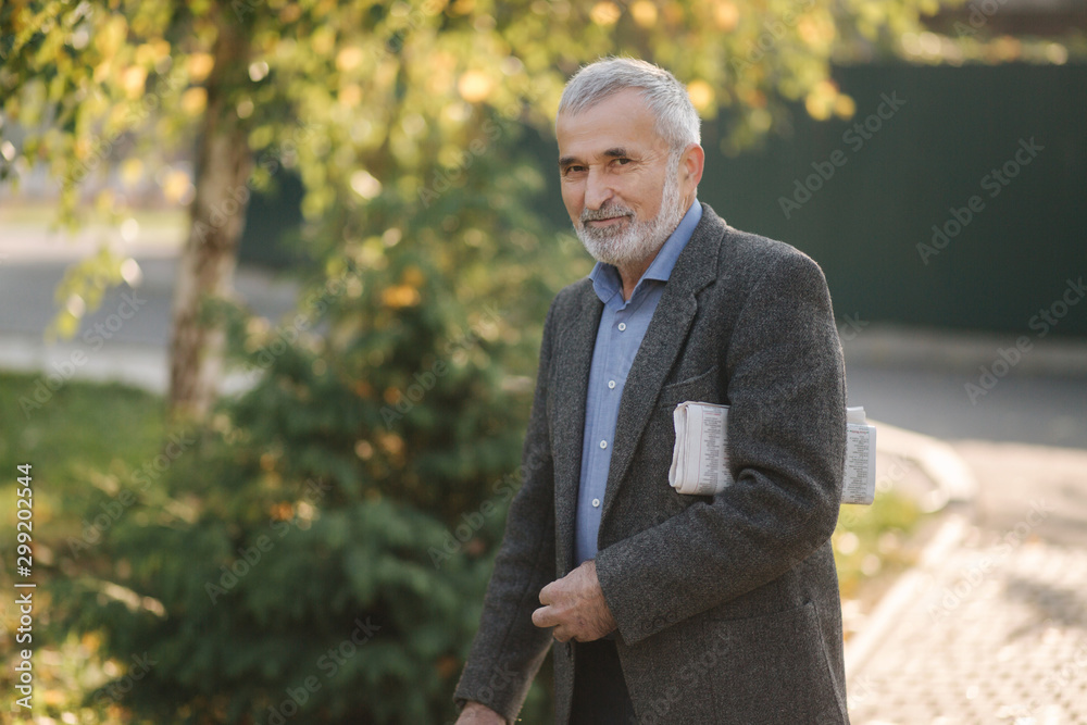 Handsome senior man walk through the park with newspaper in hands. Bearded grandpa early in the morning walking in the park Handsome senior man walk through the park with newspaper in hands. Bearded grandpa early in the morning walking in the park