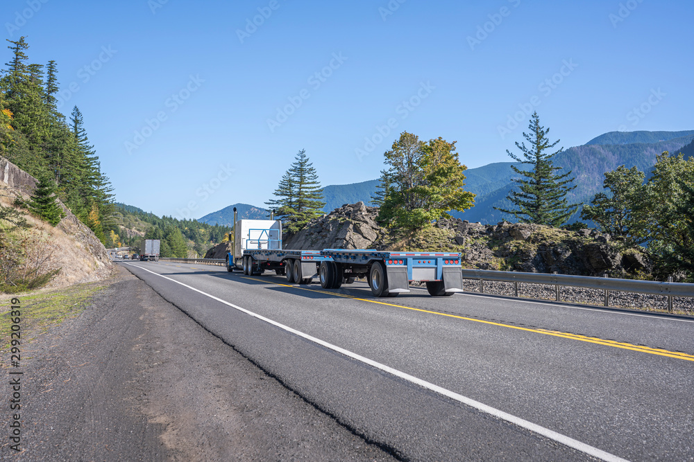Big rig blue classic semi truck transporting two empty flat bed semi trailers going to warehouse for load running on the road along the Columbia Gorge area