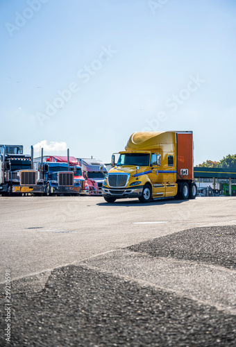 Yellow big rig semi truck with orange semi trailer driving on the truck stop driving out of a line of parked semi trucks