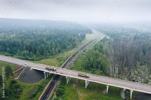 Crossing of Highway R-255 "Siberia" over road bridge with Trans-Siberian Railway in green taiga forest, smog from fires in air, aerial view. Main freight traffic routes. Krasnoyarsk Krai, Russia.