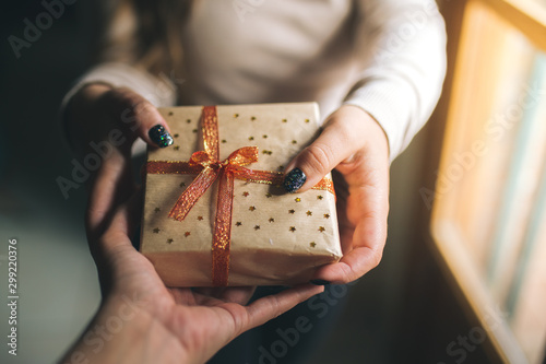 Young women with glitter nails giving a Christmas present box to his friend. Hands hold new year gift box. decorated with craft paper, red and golden ribbon and stars. Family gift and holiday concept.