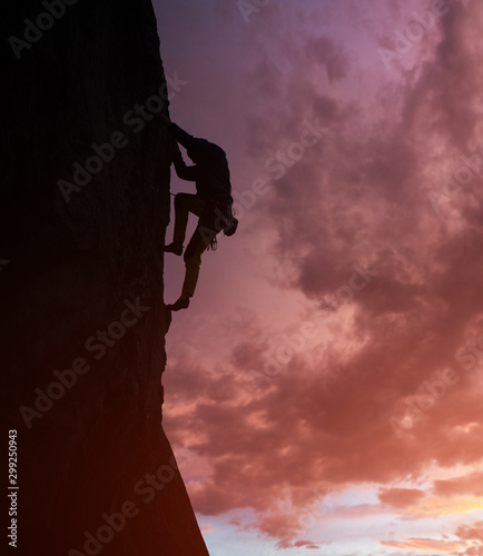 Male silhouette rock climbing and doing next step. Rock climber in action on cliff. Dark purple cloudy sky on background with copy space. Low angle view. Dangerous, extreme, endurance, victory concept