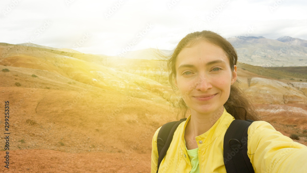 A brunette girl in a yellow jacket and backpack takes a selfie against the background of bright red sand mountains. The glare of the sun on the background of nature A brunette girl in a yellow jacket and backpack takes a selfie against the background of bright red sand mountains. The glare of the sun on the background of nature