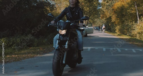 Pretty smiling European young woman driving a motorbike wearing leather jacket in forest with vibrant, colorful golden autumn leaves on sunny day. Frontal view, Slow Motion, Utrecht , Netherlands photo