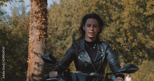 front perspective of pretty smiling European young lady driving a motorbike wearing leather jacket in forest with vibrant, colorful golden autumn leaves on sunny day. Slow Motion, Utrecht, Netherlands photo