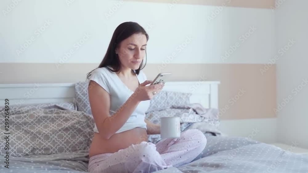 Beautiful pregnant woman is sitting on her bed in bedroom and drinking a tea. She is smiling to herself and clicking on a phone. Beautiful pregnant woman is sitting on her bed in bedroom and drinking a tea. She is smiling to herself and clicking on a phone.
