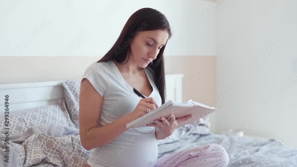 Beautiful pregnant woman is sitting on her bed in bedroom and writing something in a notebook. She is smiling to herself and caressing her belly. Beautiful pregnant woman is sitting on her bed in bedroom and writing something in a notebook. She is smiling to herself and caressing her belly.