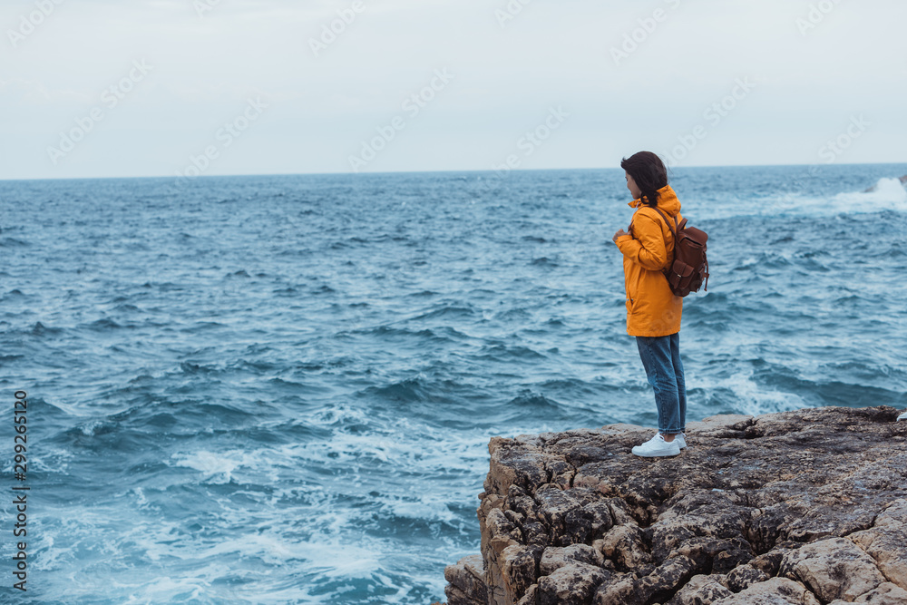 woman in yellow raincoat at the cliff enjoying sea view at stormy weather