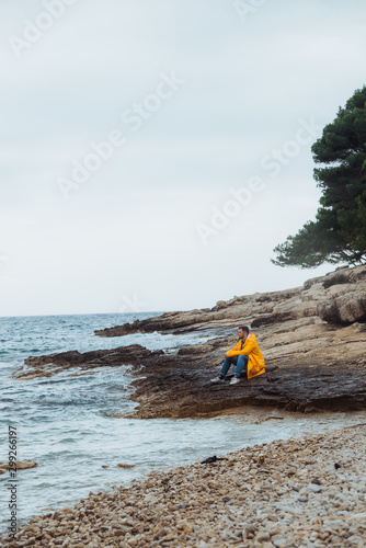 man sitting in yellow raincoat at rocky beach looking at stormy weather