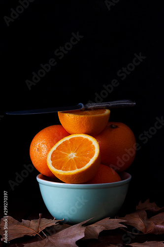 ORANGE IN BOWL WITH KNIFE NEXT TO TREE LEAVES ON DARK BACKGROUND. AUTUMN FRUIT