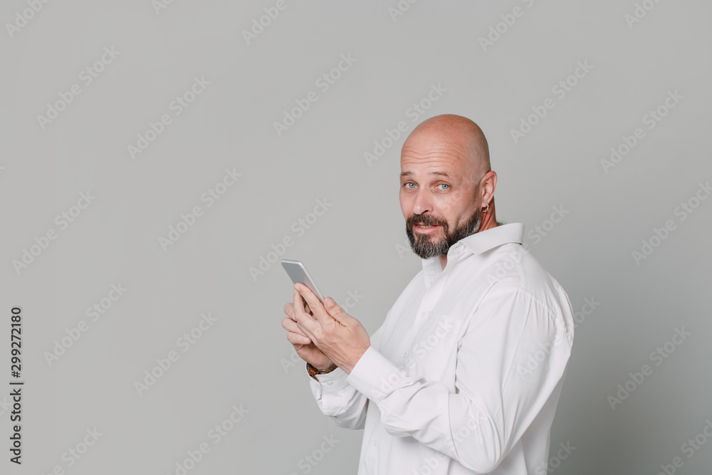 Handsome serious middle-aged man in a white shirt smiling holds a phone in his hands on a gray background with copy space Handsome serious middle-aged man in a white shirt smiling holds a phone in his hands on a gray background with copy space