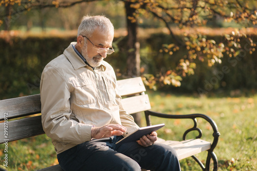Handsome elderly man sitting on the bench and use tablet for scrolling in internet. Background of autumn tree
