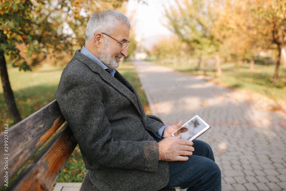 Handsome gray-haired elderly man with beautiful beard sitting on the bench and use tablet for scrolling in internet. Background of autumn tree Handsome gray-haired elderly man with beautiful beard sitting on the bench and use tablet for scrolling in internet. Background of autumn tree