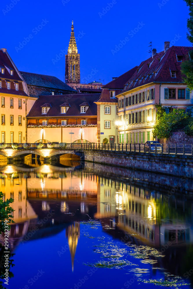 The steeple of Notre-Dame cathedral in Strasbourg, France, protruding above the roofs of the Petite France quarter illuminated at nightfall and reflecting in the still waters of the river Ill canal.