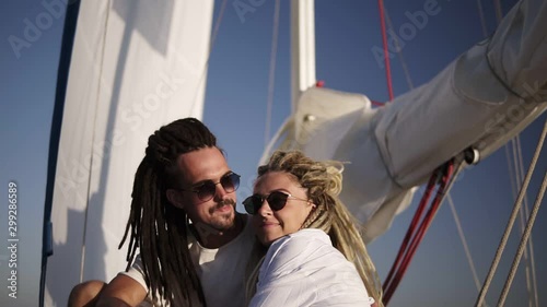 Low angle footage of stylish couple with dreadlocks in white clothes and sunglasses sitting embracing on the bow of the yacht and kissing. Loving couple spending their honeymoon on the yacht beyond photo