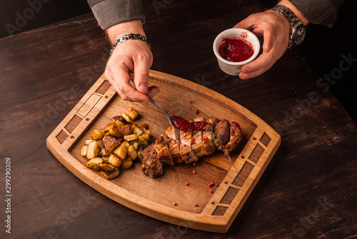 Grilled meat served with potatoes on an wooden board. Hands pouring the sauce on the meat.