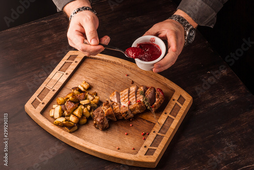 Grilled meat served with potatoes on an wooden board. Hands pouring the sauce on the meat.