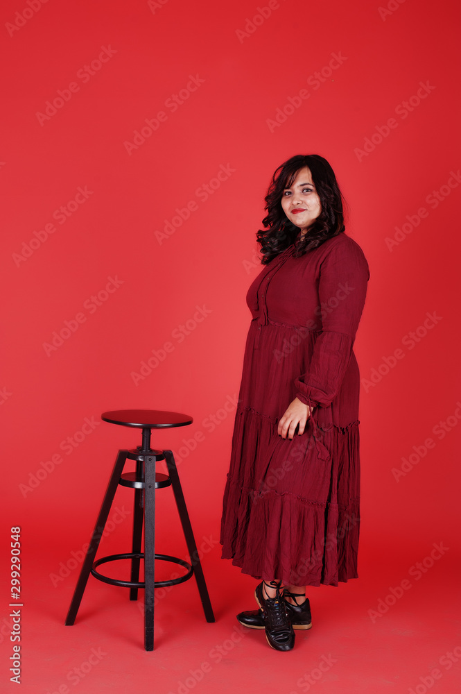 Attractive south asian woman in deep red gown dress posed at studio on pink background sitting on chair. Attractive south asian woman in deep red gown dress posed at studio on pink background sitting on chair.