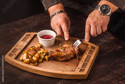 Grilled meat served with potatoes and red sauce on an wooden board. Hands cutting the meat with serving knife.