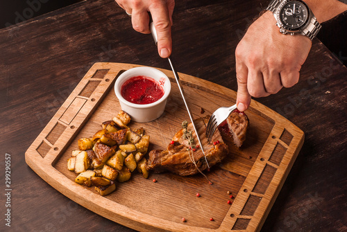 Grilled meat served with potatoes and red sauce on an wooden board. Hands cutting the meat with serving knife.