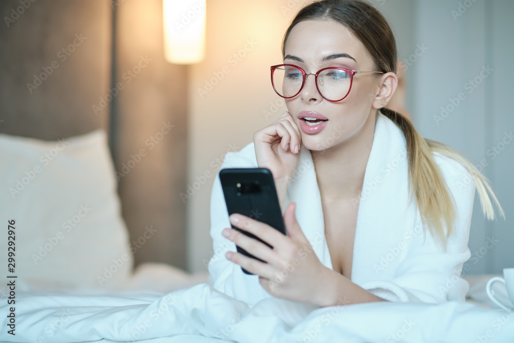Beautiful young woman in bathrobe having rest on comfortable bed room after spa procedures. She is typing message on smartphone and smiling. Lady lies on comfortable bed and uses smartphone.