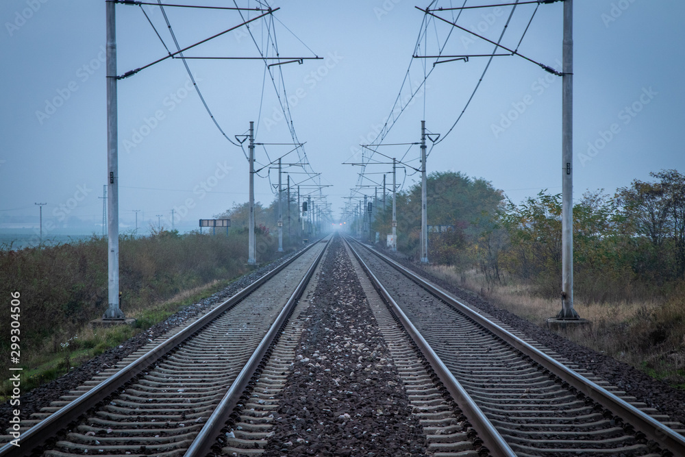 Two train tracks leading into distance. Poles with power lines overhead. Very moody and grey atmosphere. Efficient and fast way of transport.