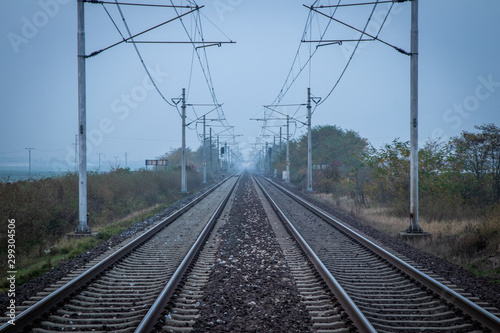 Two train tracks leading into distance. Poles with power lines overhead. Very moody and grey atmosphere. Efficient and fast way of transport.