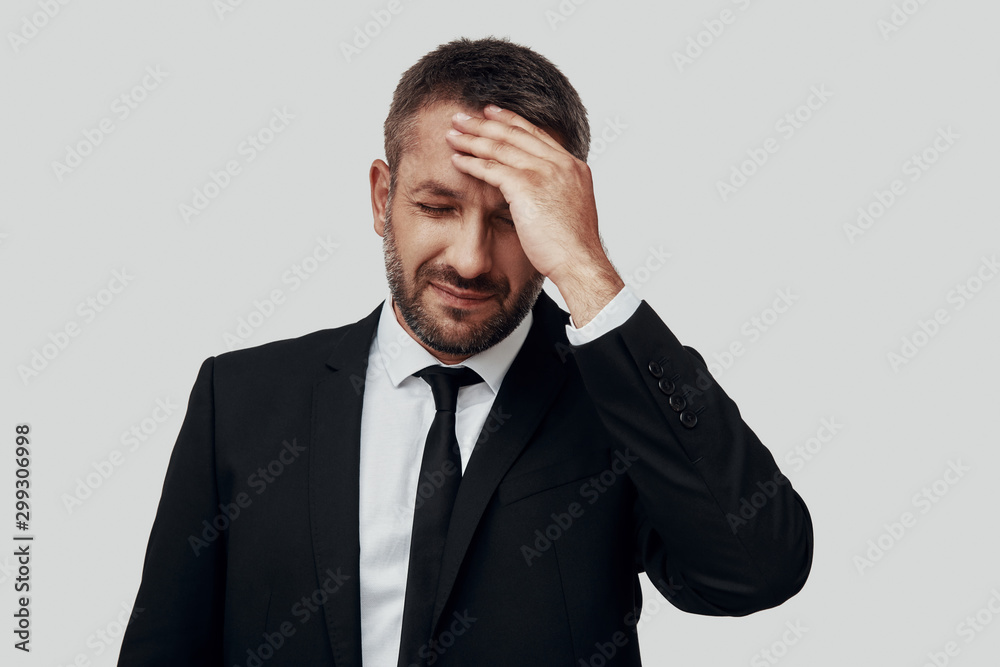 Frustrated young man in full suit suffering from headache while standing against grey background Frustrated young man in full suit suffering from headache while standing against grey background