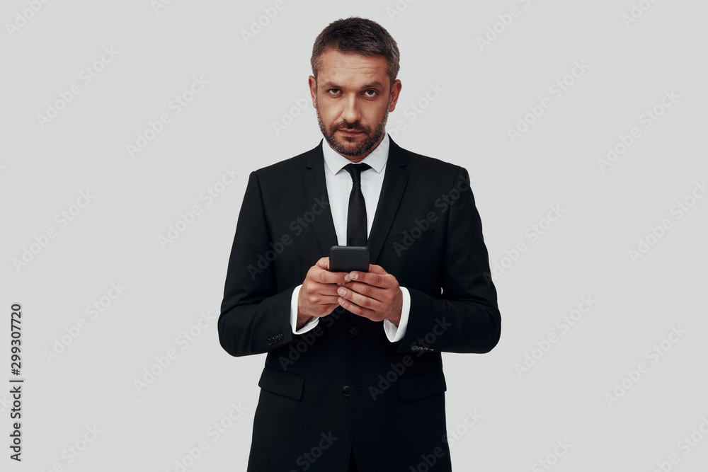Charming young man in full suit using smart phone while standing against grey background Charming young man in full suit using smart phone while standing against grey background
